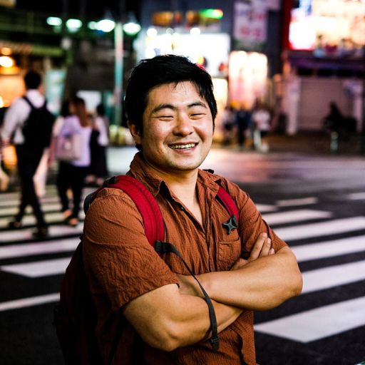 A smiling man with a red backpack standing at a crosswalk in a brightly lit Korean city at night.